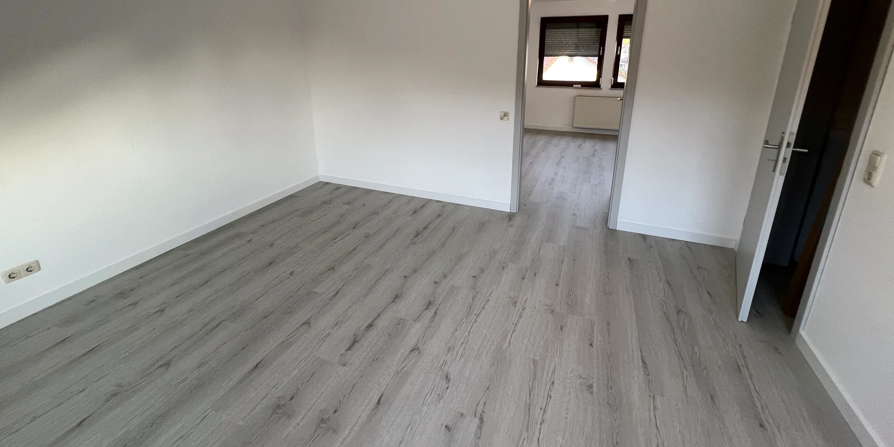 Leerer, heller Raum mit neu verlegtem Vinylboden in grauer Holzoptik und weißen Sockelleisten, der Boden läuft ins Nebenzimmer weiter