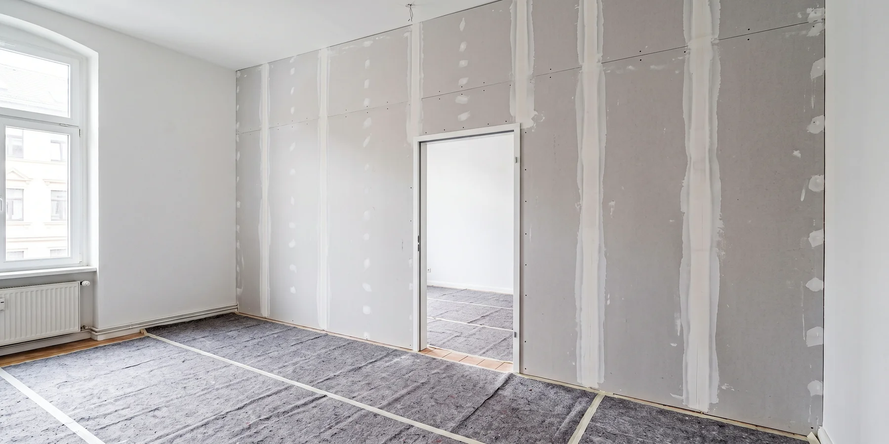 Symbolbild: Neue Trockenbauwand mit eingebauter weißer Stahlzarge in einem hellen Altbauzimmer, die senkrechten Plattenstöße frisch verspachtelt, Boden mit grauem Vlies abgedeckt